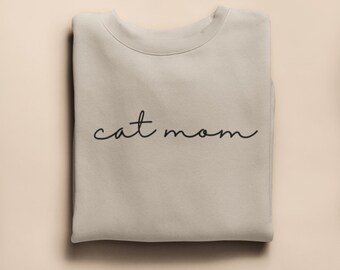 May include: A folded, pale beige sweatshirt featuring the words "cat mom" in a black cursive script. The sweatshirt is made of a soft material and is neatly folded on a light beige surface.