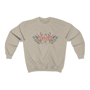 Pu&ograve; includere: Una felpa beige con la scritta "tired" in corallo, circondata da un disegno floreale con fiori rosa e bianchi e foglie verdi. La felpa ha maniche lunghe e girocollo.