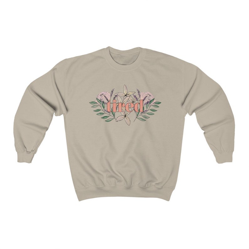 Pu&ograve; includere: Una felpa beige con la scritta "tired" in corallo, circondata da un disegno floreale con fiori rosa e bianchi e foglie verdi. La felpa ha maniche lunghe e girocollo.