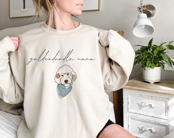 Puede incluir: Sudadera beige con la frase "goldendoodle mama" en cursiva. Debajo del texto hay un dibujo lineal de un perro goldendoodle con gafas y un pañuelo azul. La sudadera tiene mangas largas.