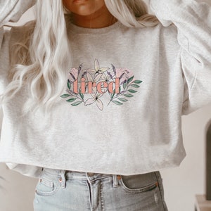 Pu&ograve; includere: Una felpa grigio chiaro con la parola "tired" in corallo, circondata da illustrazioni floreali e di foglie in rosa, viola e verde. La felpa &egrave; di stile casual.