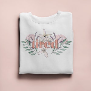 Pu&ograve; includere: Felpa bianca con la parola "tired" in pesca, circondata da un design floreale in rosa, lavanda e verde. La felpa &egrave; piegata ordinatamente su una superficie rosa chiaro.