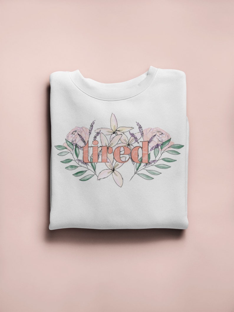 Pu&ograve; includere: Felpa bianca con la parola "tired" in pesca, circondata da un design floreale in rosa, lavanda e verde. La felpa &egrave; piegata ordinatamente su una superficie rosa chiaro.
