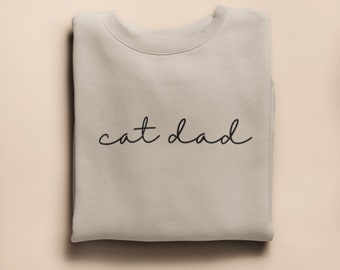 Może przedstawiać: Złożona, jasno beżowa bluza z napisem "cat dad" czarną kursywą. Bluza wykonana jest z miękkiego materiału i jest starannie złożona. Tło jest jednolite, jasno beżowe.