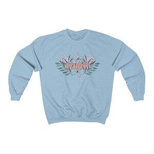 Pu&ograve; includere: Felpa azzurra con la scritta "tired" in corallo, circondata da un disegno floreale con fiori rosa e bianchi e foglie verdi. La felpa ha maniche lunghe e girocollo.