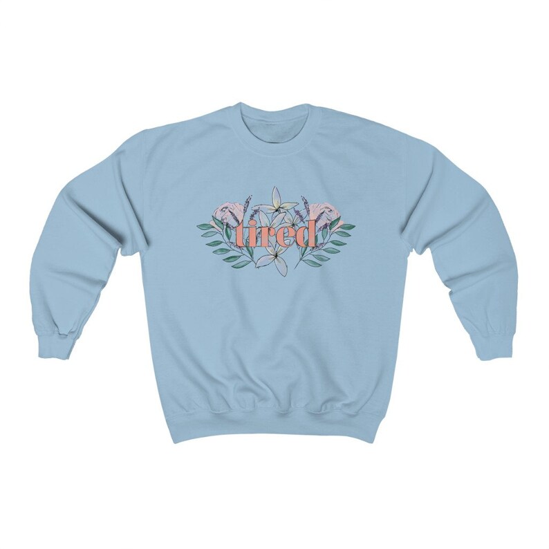 Pu&ograve; includere: Felpa azzurra con la scritta "tired" in corallo, circondata da un disegno floreale con fiori rosa e bianchi e foglie verdi. La felpa ha maniche lunghe e girocollo.