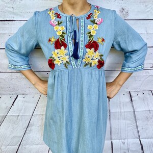 Op de afbeelding: Een lichtblauwe denimjurk met geborduurde bloemenmotieven in rood, geel en roze. De jurk heeft een striknek met een kwast.