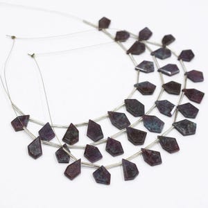 May include: A necklace with multiple strands of dark purple, geometric beads. The beads have a faceted appearance and are strung on thin, silver wire. The necklace is displayed against a white background.