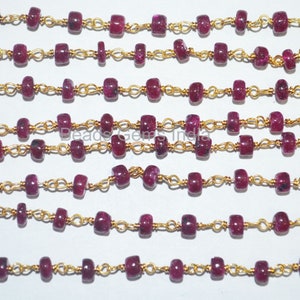 Könnte beinhalten: Eine Strähne aus rubinroten Perlen, die auf einer goldenen Kette aufgefädelt sind. Die Perlen sind facettiert und haben einen leichten Glanz. Die Kette besteht aus kleinen, filigranen Gliedern.