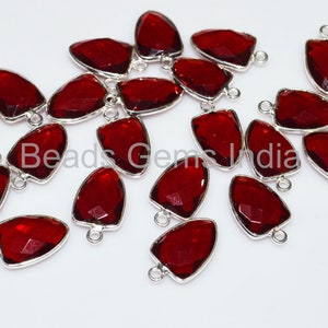 Puede incluir: Una colección de colgantes de piedras preciosas rojo oscuro, facetados, en forma de escudo, con bordes de metal plateado y pequeños bucles para sujetar a la joyería. Los colgantes están dispersos sobre una superficie blanca. El texto "Beads Gems India" es parcialmente visible.