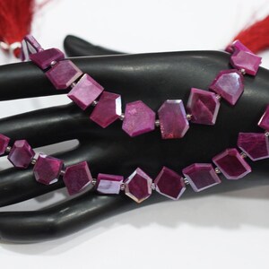 May include: Two strands of faceted, deep magenta gemstone beads. The beads are irregularly shaped with flat surfaces and connected by small silver-coloured links. Red tassels are visible at the ends of the strands. The beads are displayed on a black surface.