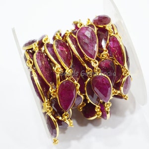 Puede incluir: Una hebra de cuentas de rubí con detalles dorados. Las cuentas son facetadas y tienen forma de lágrima. La hebra está enrollada sobre un fondo blanco.