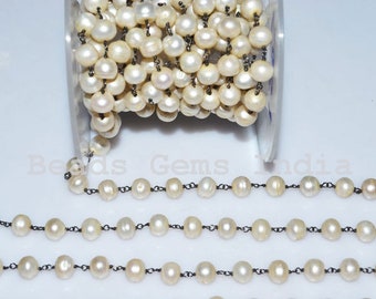Op de afbeelding: Een streng witte zoetwaterparels geregen aan een zwarte ketting. De parels zijn rond en hebben een lichte iriserende glans. De ketting is gemaakt van kleine, delicate schakels.