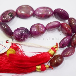 Può includere: Una collana di 18 perle di rubino ovali di colore rosa intenso e viola. Le perle sono infilate su un cordoncino bianco con nappe rosse ad ogni estremità. La collana è etichettata "Beads Gems India".