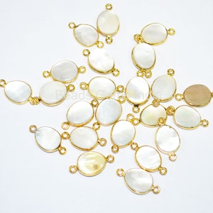 May include: A group of white oval-shaped shell beads with gold-toned metal connector loops. The beads are scattered on a white surface.
