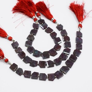 May include: Three strands of faceted, dark purple gemstone beads with red tassel accents. The beads are a geometric shape, and the tassels are attached with silver-coloured metal. The beads are displayed on a white background.
