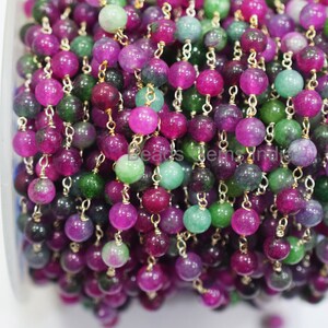 May include: A close-up of a spool of green, purple, and pink gemstone beads strung on a gold chain. The beads are round and have a smooth, polished finish.