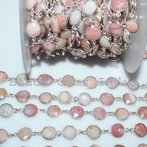 May include: A silver chain with pink and white gemstone beads. The chain is coiled up on a white surface. The beads are oval shaped and have a faceted finish.