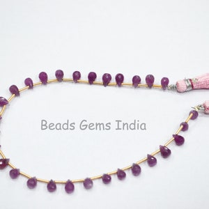 May include: A strand of purple gemstone beads strung on a gold wire with pink tassels on each end. The beads are teardrop shaped and have a faceted finish. The text "Beads Gems India" is visible in the center of the image.