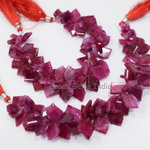 以下が含まれることがあります： 不規則なファセットカットが施されたピンクのルビーのビーズのネックレス。ビーズは赤いタッセルが両端に付いたコードに糸に通されています。ネックレスは白い表面に置かれています。  "Beads Gems India"が表面に印刷されています。