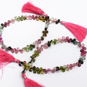 Puede incluir: Hilos de cuentas de piedras preciosas de turmalina coloridas con borlas rosas brillantes. Las cuentas son una mezcla de verde, rosa y transparente, dispuestas en forma de lágrima. Las borlas son de un fucsia vibrante.