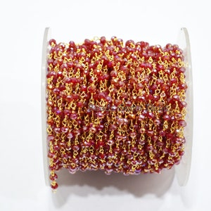 Puede incluir: Un carrete de cadena dorada con cuentas de cristal facetadas rojas. La cadena está enrollada alrededor de un carrete de plástico transparente.