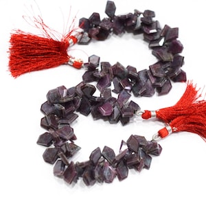 以下が含まれることがあります： ファセットカットの濃い紫色の宝石ビーズの2つのストランド。各ストランドは、両端に鮮やかな赤いタッセルで飾られています。ビーズは白い背景に配置されています。