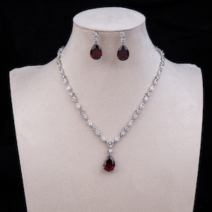 May include: A silver necklace with a teardrop-shaped red gemstone pendant and matching earrings. The necklace is adorned with small, clear stones.