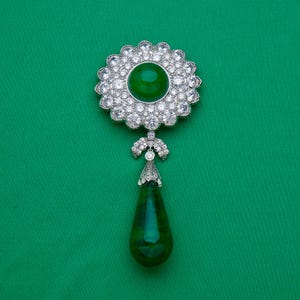 May include: A silver-toned brooch with a large, round, emerald-green center stone surrounded by a halo of clear, sparkling crystals. A smaller, teardrop-shaped green stone hangs below, connected by a decorative silver element. The background is a solid, vibrant green.
