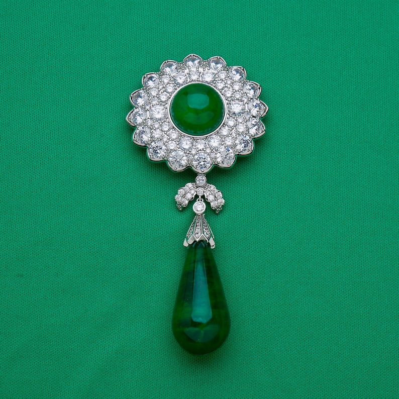 May include: A silver-toned brooch with a large, round, emerald-green center stone surrounded by a halo of clear, sparkling crystals. A smaller, teardrop-shaped green stone hangs below, connected by a decorative silver element. The background is a solid, vibrant green.