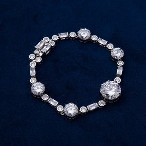 May include: A silver bracelet with multiple round and rectangular clear gemstones. The bracelet features a series of large, round stones interspersed with smaller, rectangular and round stones. The bracelet is displayed on a dark blue surface.