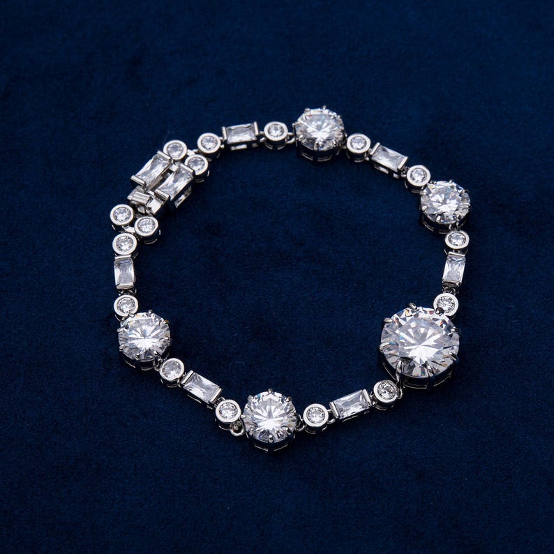 May include: A silver bracelet with multiple round and rectangular clear gemstones. The bracelet features a series of large, round stones interspersed with smaller, rectangular and round stones. The bracelet is displayed on a dark blue surface.