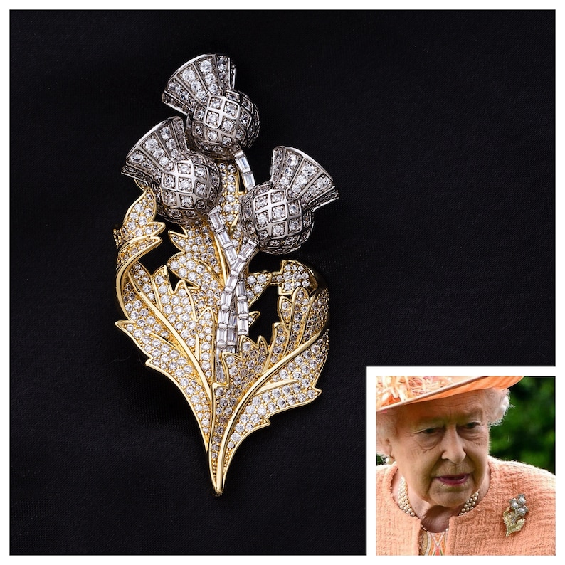 May include: A silver and gold brooch featuring a cluster of three thistle flowers with clear rhinestones. The brooch is a traditional Scottish symbol.