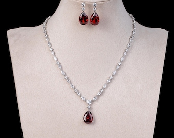 May include: A silver-toned necklace and earring set displayed on a mannequin. The necklace and earrings feature teardrop-shaped, deep red gemstones. The necklace has a series of smaller, clear stones. The earrings are a matching pair.