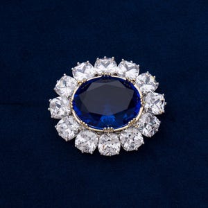 May include: A detailed close-up of a brooch with a large, oval, deep blue gemstone at the center, surrounded by a halo of square, clear gemstones. The setting is gold-toned, and the background is a dark blue.