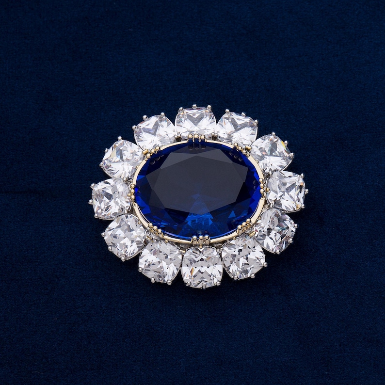 May include: A detailed close-up of a brooch with a large, oval, deep blue gemstone at the center, surrounded by a halo of square, clear gemstones. The setting is gold-toned, and the background is a dark blue.