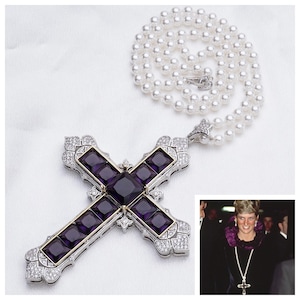 Könnte beinhalten: Ein silberner und goldener Kreuzanhänger mit violetten Edelsteinen und einer Perlenkette. Das Kreuz ist mit klaren Steinen besetzt und hat ein detailliertes Design.