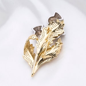 May include: A gold and silver brooch in the shape of a thistle flower. The brooch is covered in small, sparkling stones.