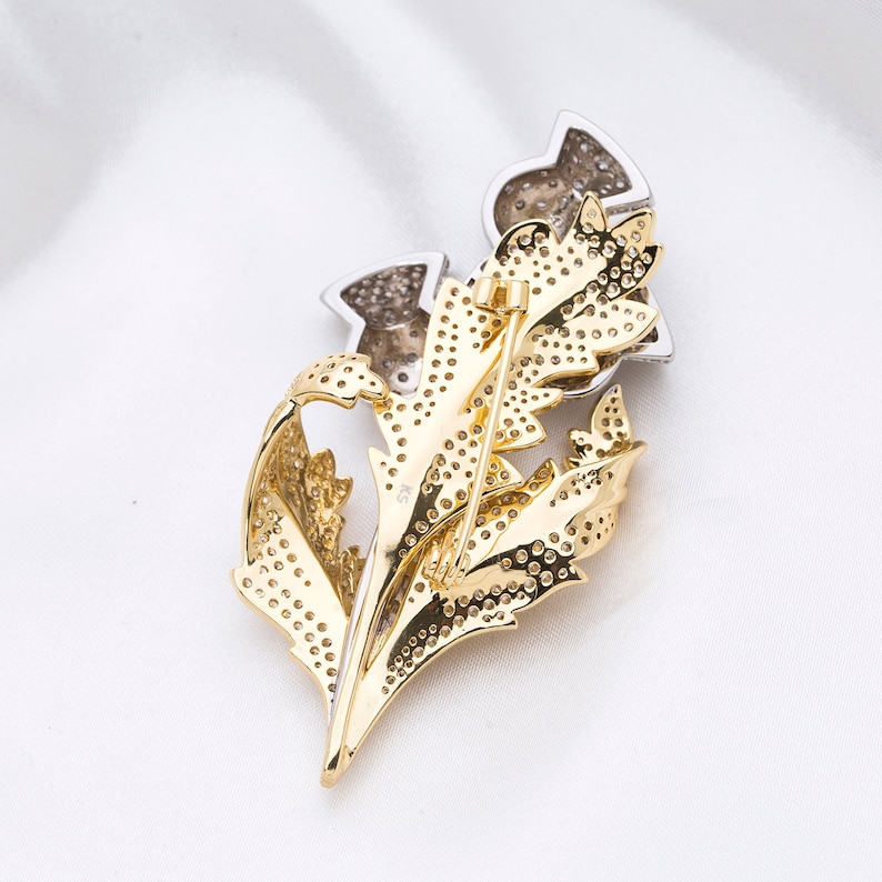 May include: A gold and silver brooch in the shape of a thistle flower. The brooch is covered in small, sparkling stones.