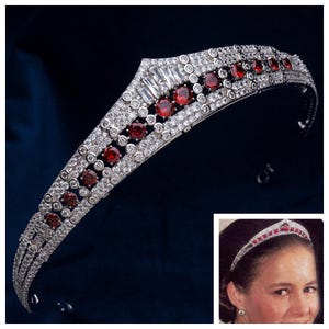 May include: A silver-toned tiara featuring a central peak design adorned with numerous small clear round crystals, baguette-cut clear crystals at the apex, and a series of deep red round stones arranged in a linear pattern along the band.