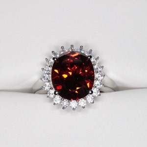 May include: A silver ring with a large, oval-shaped, red gemstone surrounded by a halo of smaller, white gemstones.
