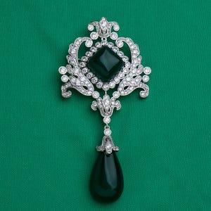 May include: A silver-toned brooch featuring a central square dark green stone surrounded by clear crystals. The ornate metalwork includes intricate scroll patterns and a dangling teardrop-shaped dark green stone at the bottom, all accented with small clear crystals.
