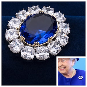 May include: A dazzling brooch featuring a large, oval, deep blue gemstone at its center, surrounded by a halo of clear, square-cut crystals. The setting is gold-toned, and the brooch is set against a dark blue background.