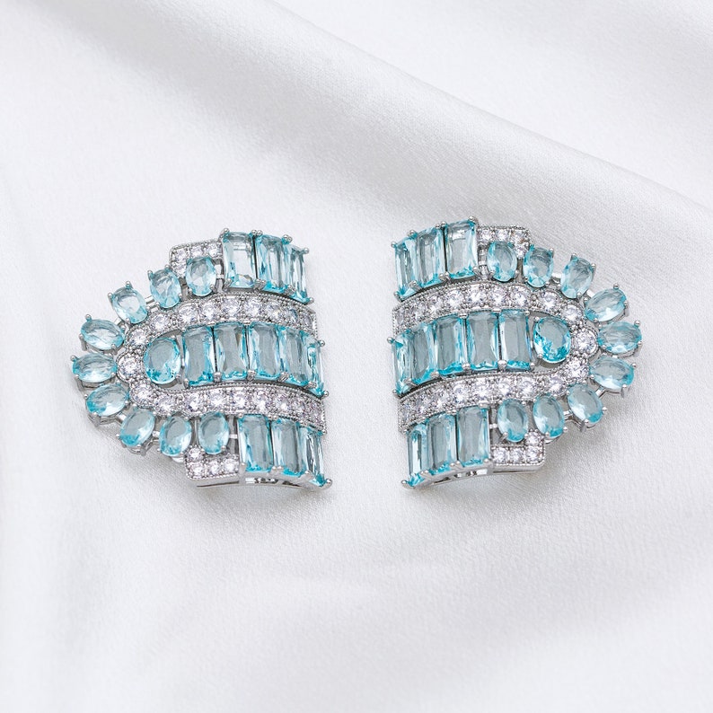 K&ouml;nnte beinhalten: Ein Paar silberne Ohrringe mit blauen und klaren Kristallsteinen. Die Ohrringe sind f&auml;cherf&ouml;rmig und haben ein geometrisches Design.