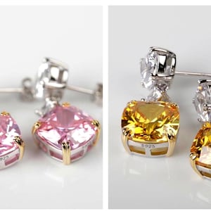 Könnte beinhalten: Ein Paar Hängeohrringe mit einem großen quadratischen rosafarbenen Edelstein und einem kleineren runden, klaren Edelstein. Die Ohrringe sind aus Silber und haben einen goldenen Rand.