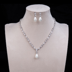 Puede incluir: Un collar de plata con un colgante de perla en forma de lágrima y pendientes a juego. El collar está adornado con pequeños diamantes de imitación transparentes.