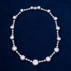 May include: A dazzling necklace featuring a series of large, round, clear gemstones interspersed with smaller, rectangular and round stones. The necklace is set in a silver or platinum metal and rests against a dark blue background.