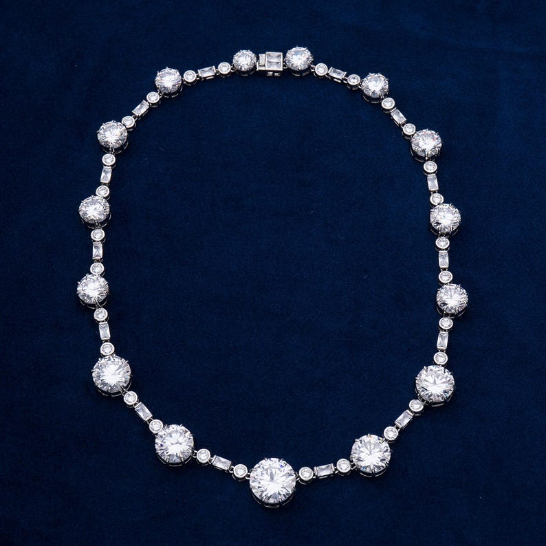 May include: A dazzling necklace featuring a series of large, round, clear gemstones interspersed with smaller, rectangular and round stones. The necklace is set in a silver or platinum metal and rests against a dark blue background.