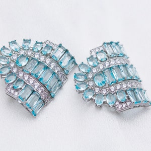 K&ouml;nnte beinhalten: Ein Paar silberne Ohrringe mit blauen und wei&szlig;en Edelsteinen. Die Ohrringe sind f&auml;cherf&ouml;rmig und haben ein filigranes Design.