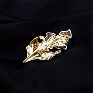 May include: A gold-tone leaf-shaped brooch with clear and black rhinestones. The brooch is set on a black fabric background.